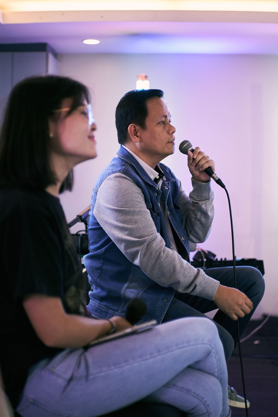Two adults singing together into handheld microphones at a karaoke session under warm stage lighting