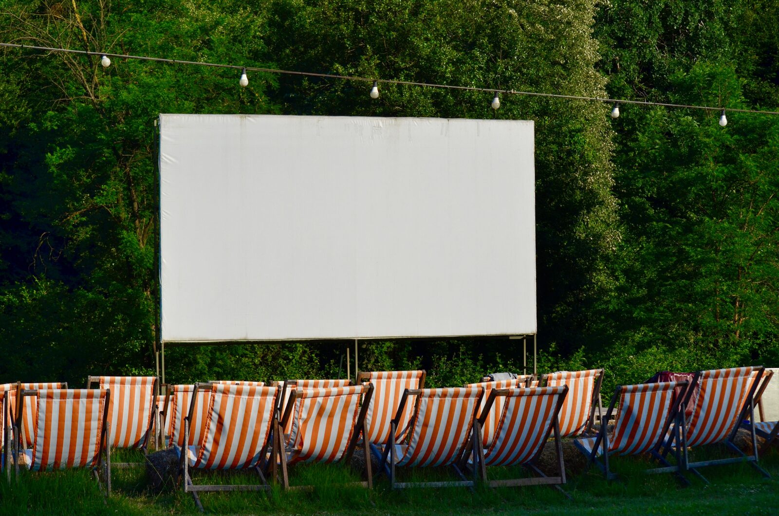 An outdoor movie setup at dusk with a large blank screen, striped deck chairs and string lights